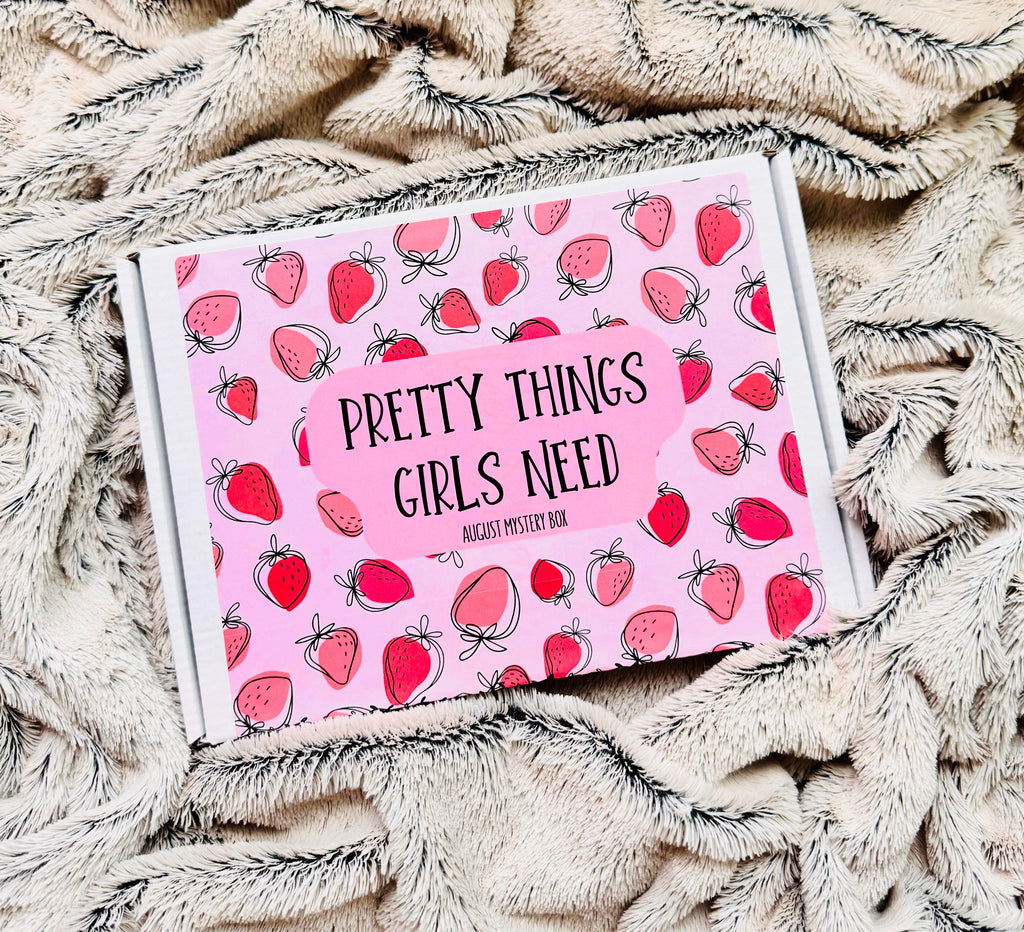 A boxed product labeled 'PRETTY THINGS GIRLS NEED' with a pink strawberry pattern, placed on a soft beige textured blanket.
